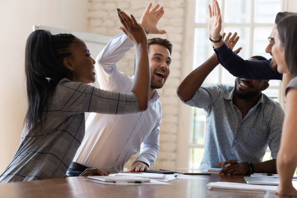 Multiracial euphoric business team people give high five at office table, happy excited diverse work group engaged in teambuilding celebrate corporate success win partnership power teamwork concept